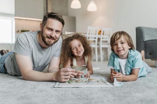 Father and two children smiling while solving a puzzle together on a soft carpet indoors.