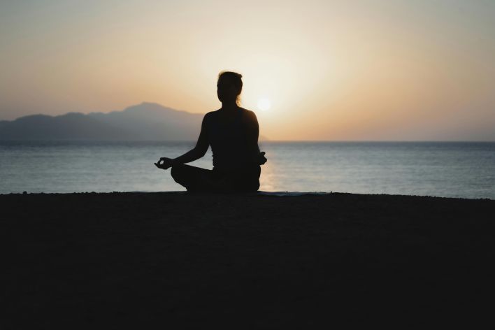 A woman practices meditation with a tranquil ocean sunset backdrop, embodying peace and relaxation.