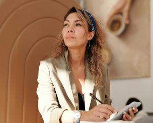 Confident woman in blazer taking notes with pen and smartphone in cafe.