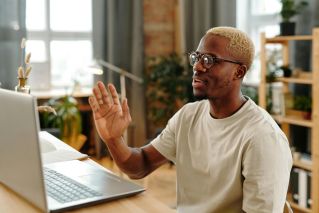 Smiling man in glasses waves during a video call at home. Well-lit room with plants.