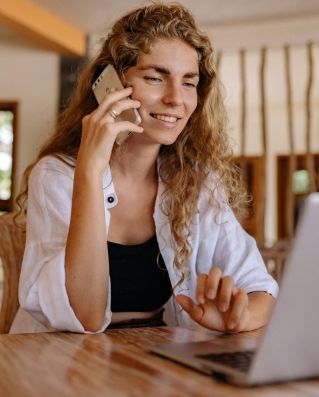 Smiling woman working remotely with a laptop and phone in a cozy setting.