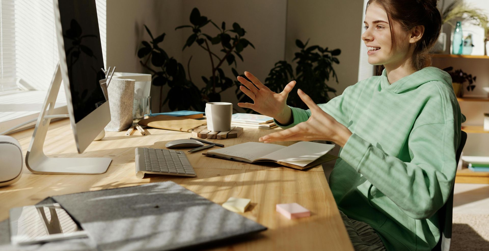 A woman in a green hoodie having a video call at her home office desk.