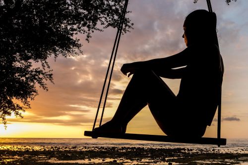 A serene sunset silhouette of a woman sitting on a swing by the seashore, evoking tranquility and reflection.