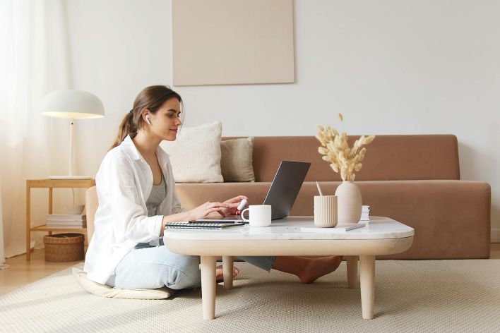 A woman uses a laptop at a coffee table in a cozy living room, embodying a relaxed work-from-home lifestyle.