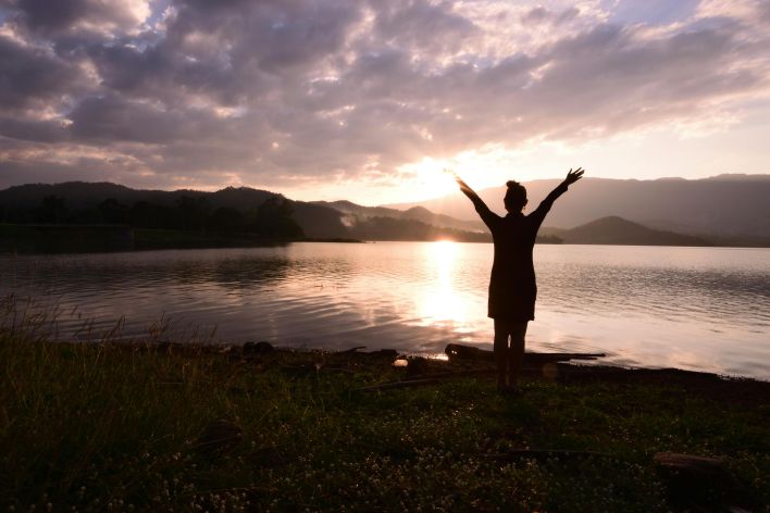 Silhouette of a woman with raised arms by a serene lake at sunset with dramatic clouds.