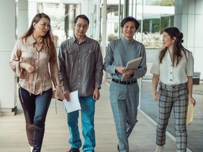 Four diverse adults walking and conversing in a modern office corridor, holding documents.