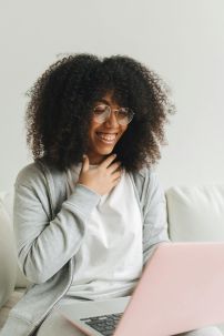 A joyful woman with curly hair uses a laptop indoors, wearing glasses and a gray hoodie.