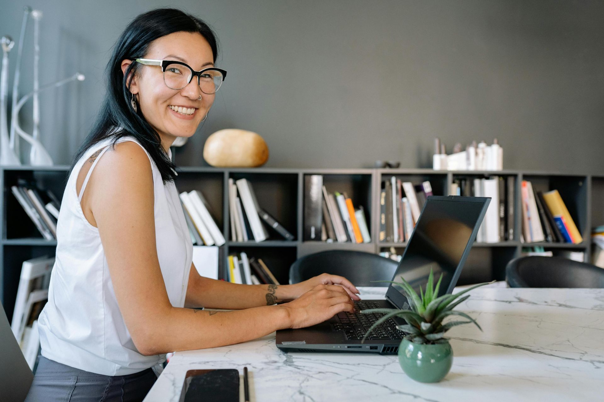 Cheerful businesswoman using laptop at a desk in a stylish office setup with bookshelves.