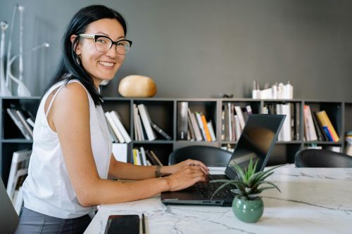 Cheerful businesswoman using laptop at a desk in a stylish office setup with bookshelves.