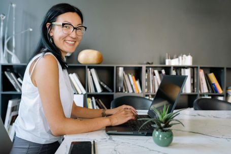 Cheerful businesswoman using laptop at a desk in a stylish office setup with bookshelves.